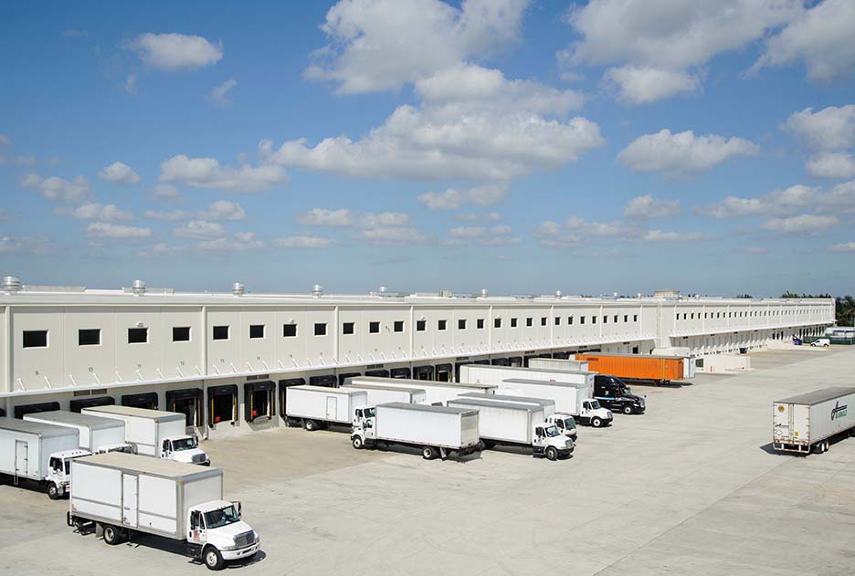 Exterior warehouse shot of Prologis Park Beacon Lakes 13 in Miami, Florida loading dock with white semi trucks driving in and out and orange truck parked